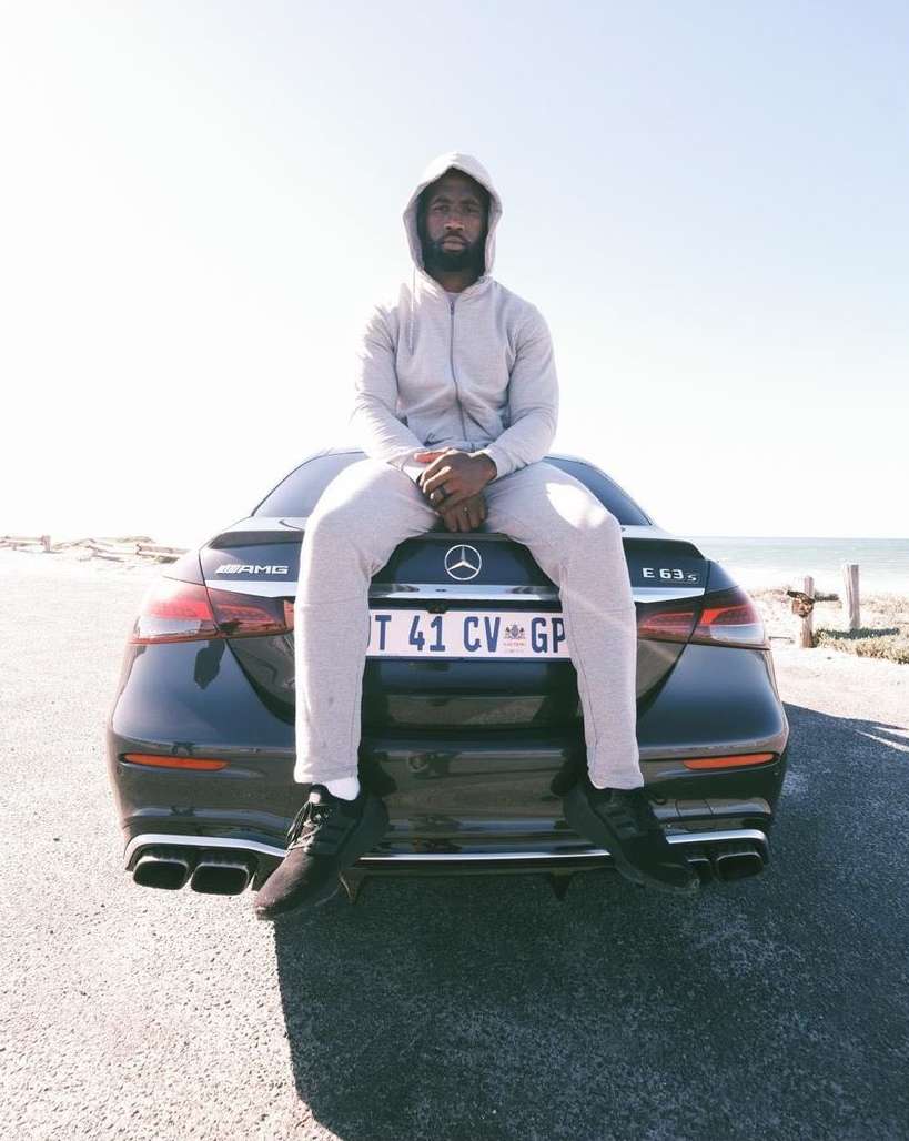 Man in gray tracksuit sits on the trunk of a black Mercedes AMG car on a road, under bright daylight, against a coastal landscape.