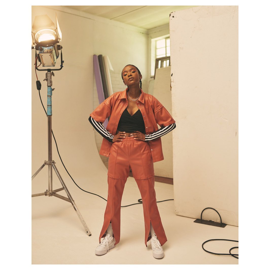 A Black female model in an orange Adidas suit poses confidently in a photo studio with lighting equipment visible in the shot.
