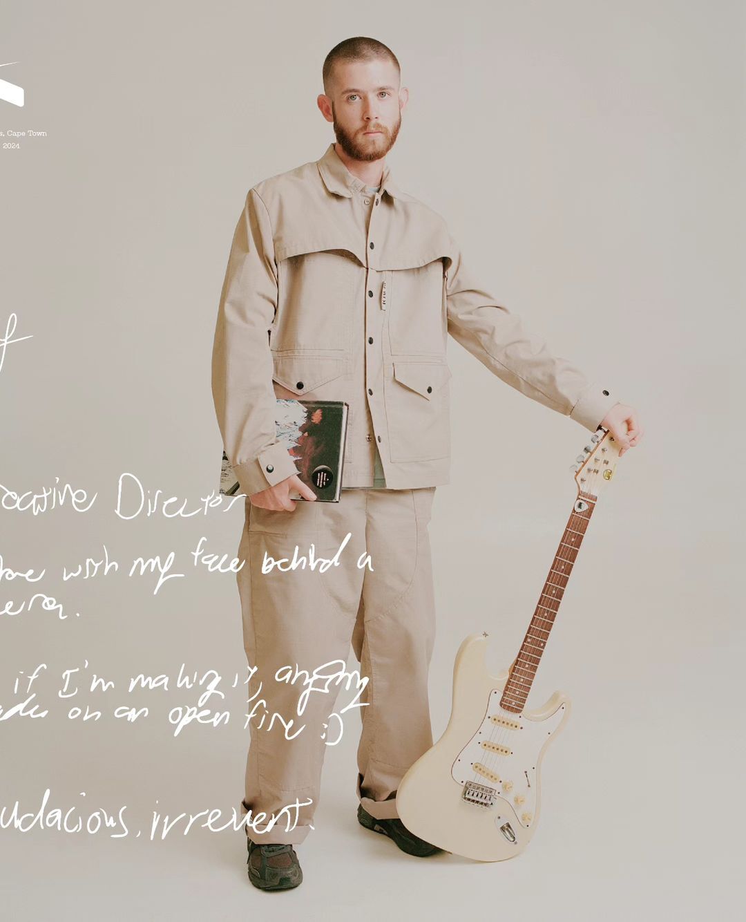 Standing adult male in casual outfit, holding guitar and book, looking at the viewer in a photo studio with a neutral backdrop.