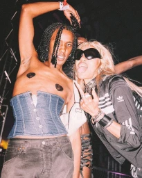 Two individuals posing closely in a dimly lit venue, wearing a denim corset top and a black jacket, captured by Dune Tilley.