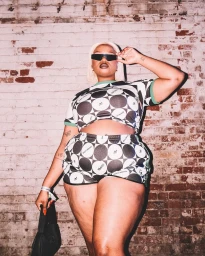 Plus-size model in a patterned two-piece outfit wearing sunglasses in front of an exposed brick wall.