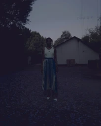 A dark, moody portrait of a Black woman standing outside a building at dusk, wearing a white tank top and pleated skirt.