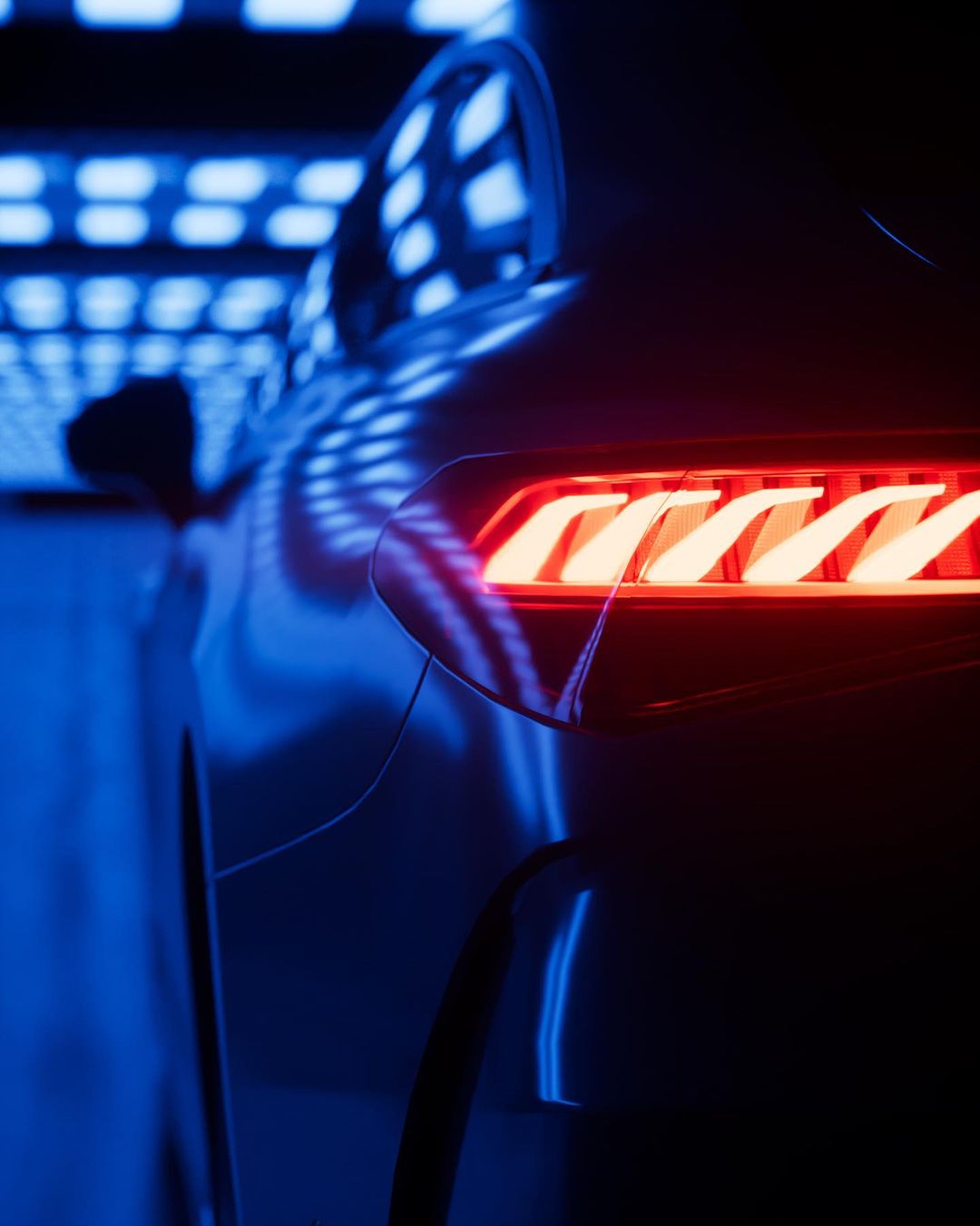 Close-up view of a dark grey car's tail light with a red and orange glow, set against a backdrop of blue and white patterned lights.