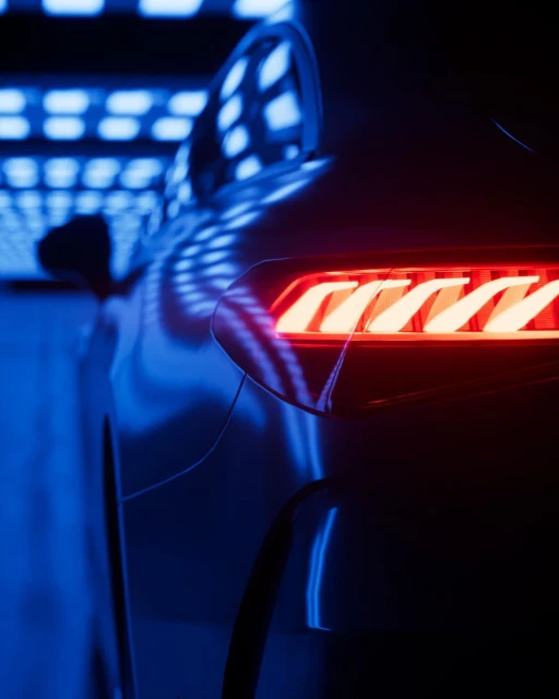 Close-up view of a dark grey car's tail light with a red and orange glow, set against a backdrop of blue and white patterned lights.