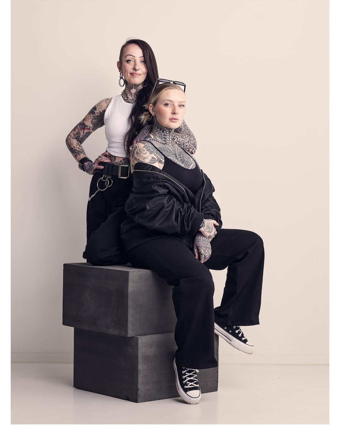 Two tattooed women pose in a studio setting. One woman sits on concrete blocks as the other stands behind her.