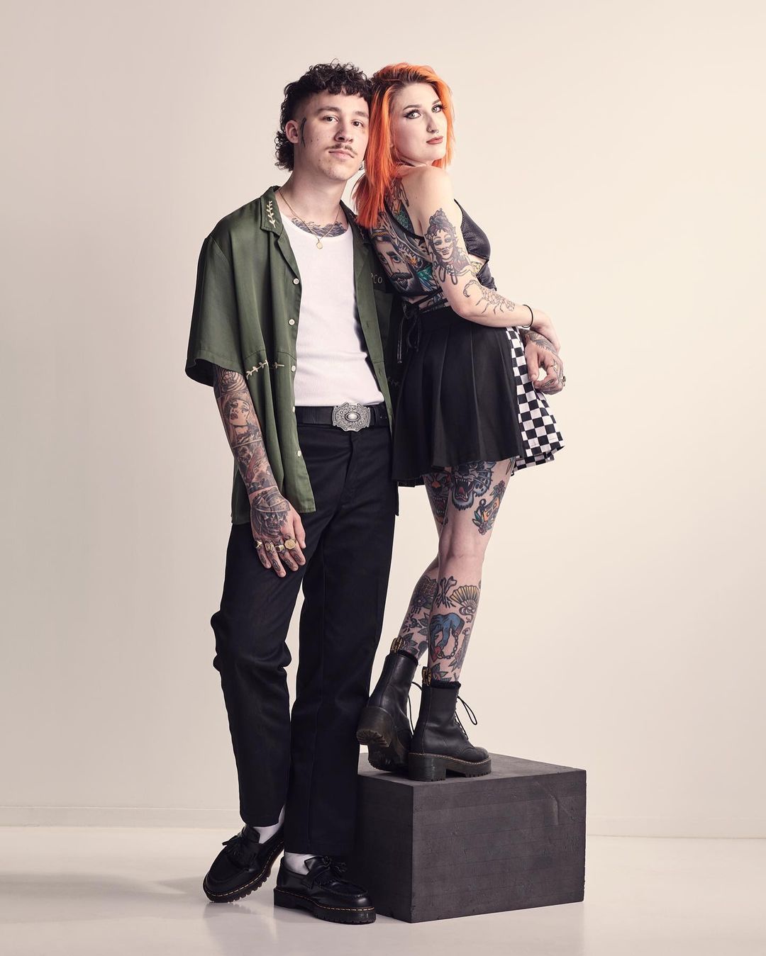 A stylish couple poses in a photo studio, showcasing fashion and tattoos. The man wears a green shirt, while the woman sports a black skirt.