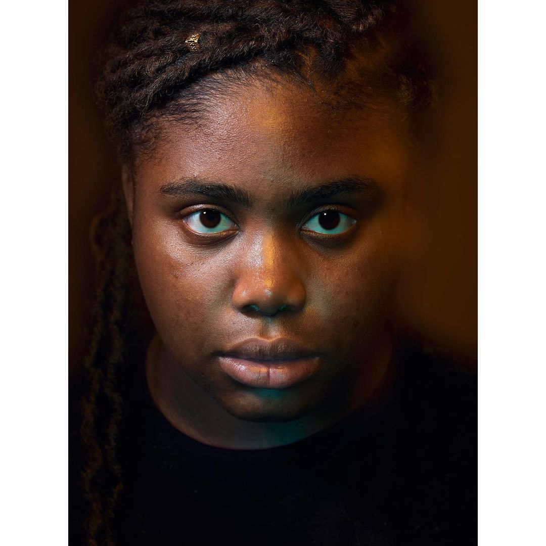 Close-up portrait of a young Black woman with dreadlocks looking directly at the camera with a neutral expression, under studio lighting.