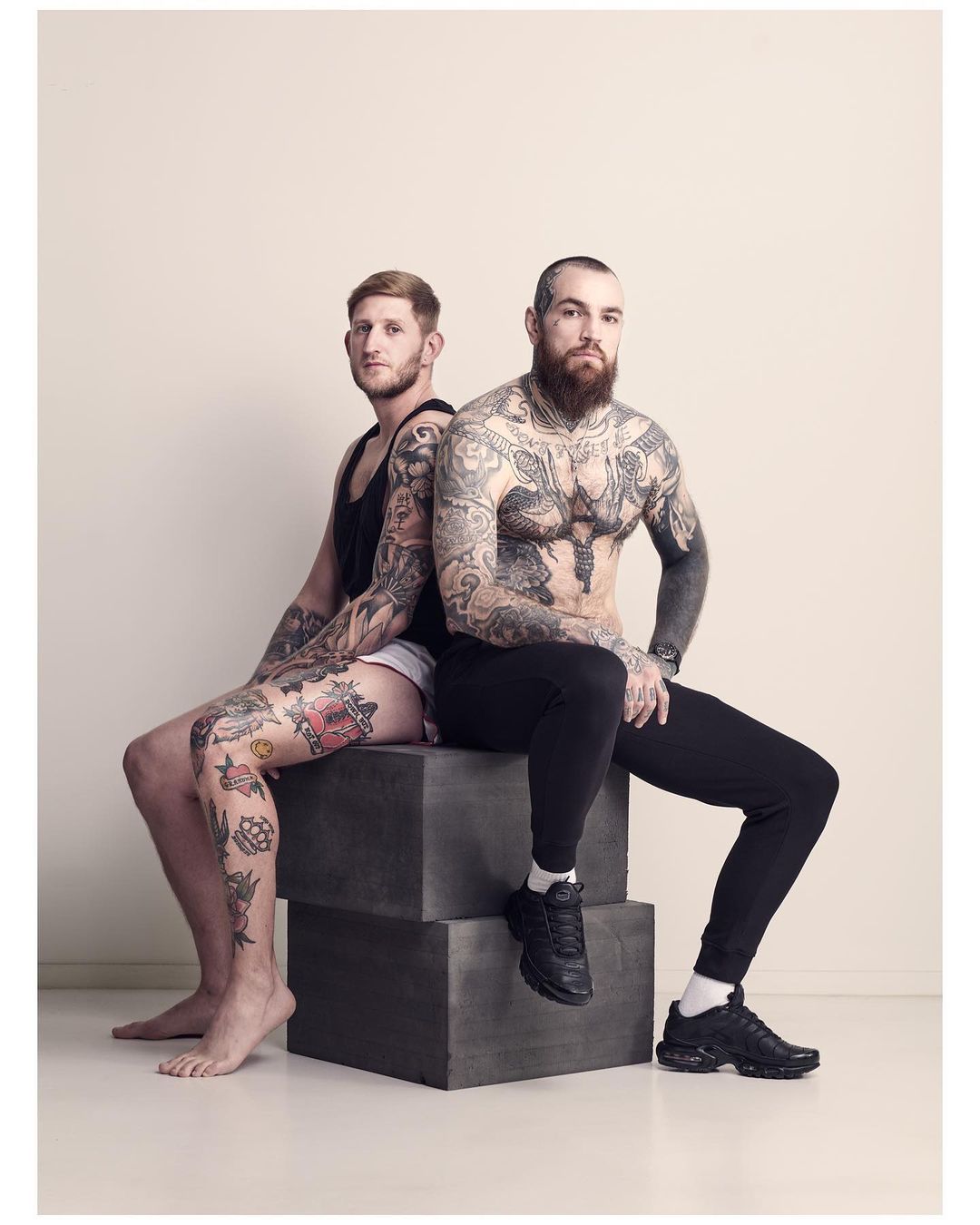 Two tattooed men sit back-to-back on gray blocks in a photo studio. One has a beard, while the other has light red hair.