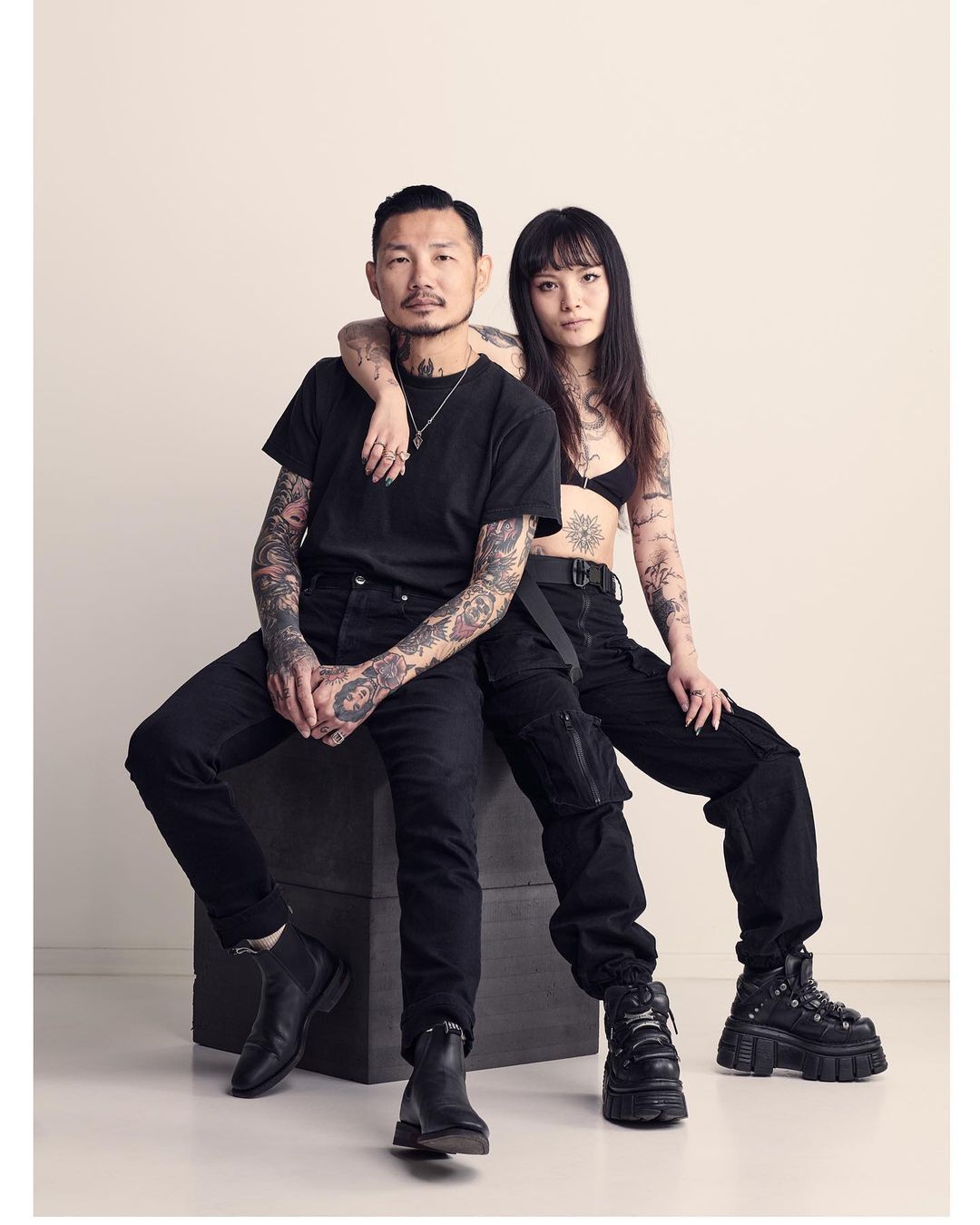 Tattooed couple in black clothing sitting indoors. Asian man with arm around woman, dark outfits, edgy fashion, and studio lighting.