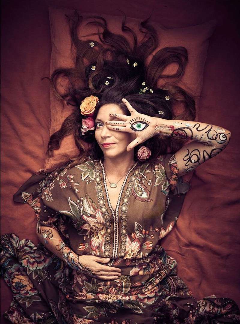 A woman lies on an orange sheet wearing a floral dress, hand painted with an eye and flowers in her hair, top-down perspective.