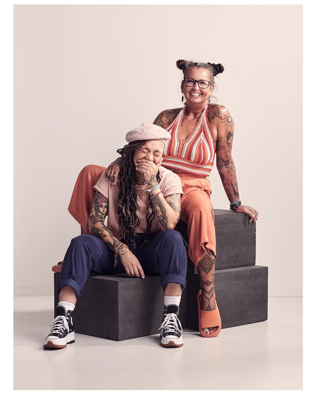Two women pose together indoors: one laughing in jeans and sneakers, the other smiling in orange halter top and pants, seated on blocks.