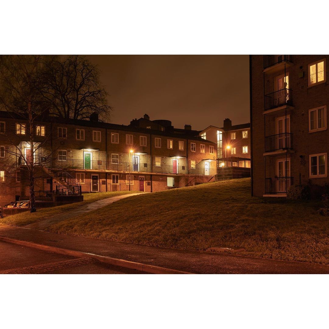 Exterior shot of apartment buildings with lighted windows at night, with green grass and a dimly lit street in the foreground.