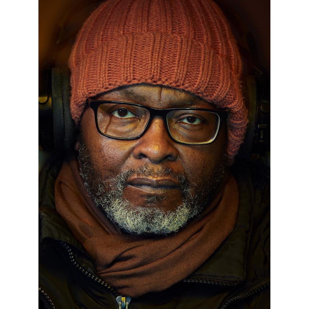 Close-up portrait of an adult male wearing a red knit hat, dark-rimmed glasses, and a brown scarf in an indoor setting with soft lighting.
