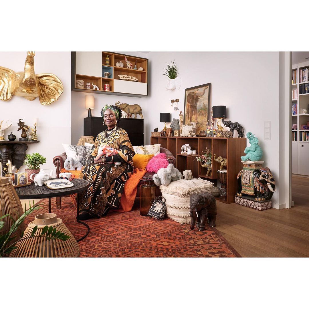 Woman in patterned dress sits in living room surrounded by elephant figurines and decor, creating a cozy, eclectic interior setting.