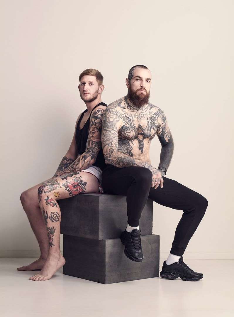Two heavily tattooed men pose together sitting on stacked grey blocks in an indoor studio with neutral background and studio lighting.