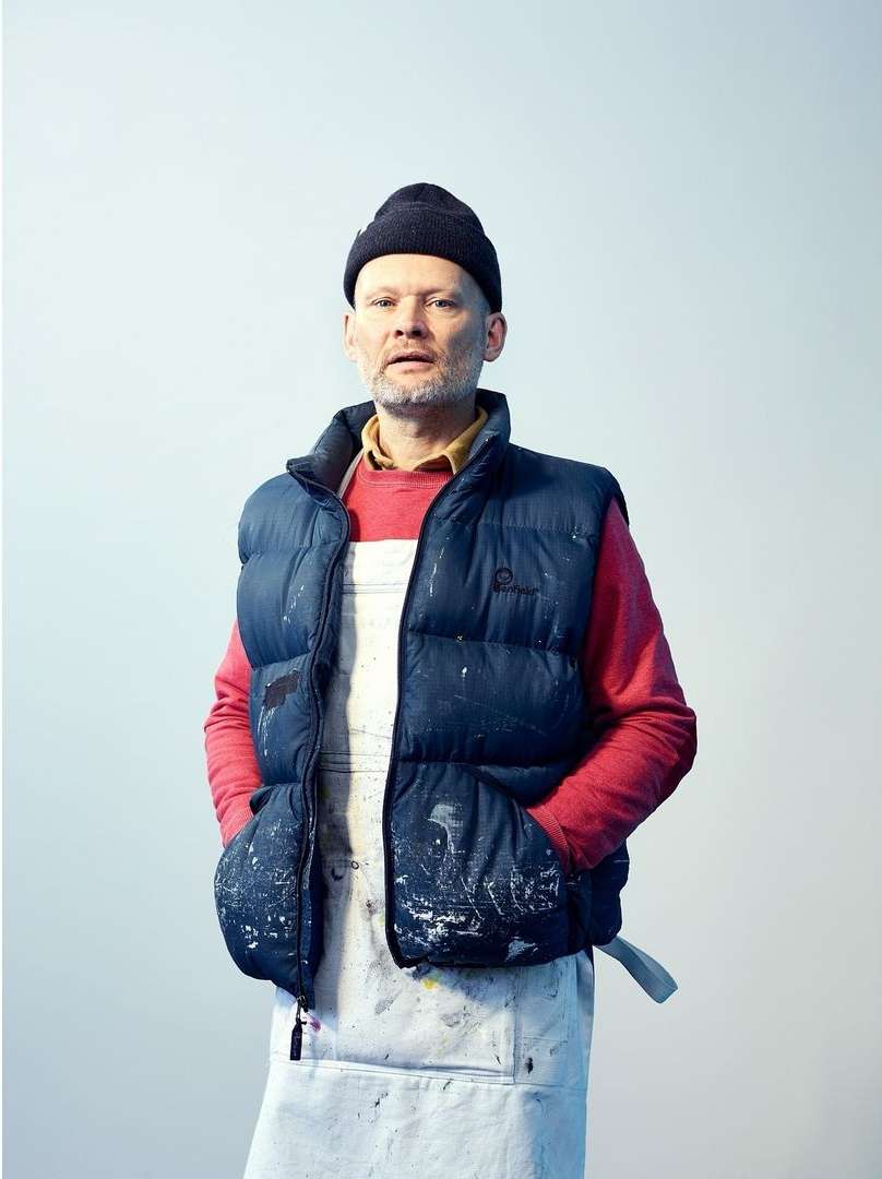 Full shot of an adult male wearing a beanie, puffer vest, and artist's apron, standing indoors against a neutral backdrop, looking at the camera.