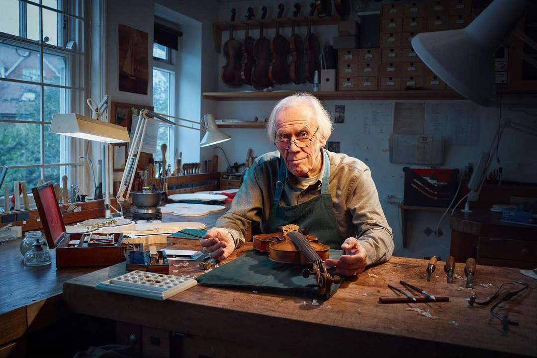 Elderly man working on a violin in his workshop with tools and instruments. Wood table, natural light from window, indoor environment.