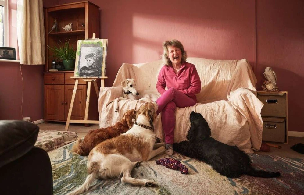 Laughing woman sitting on a couch in a living room with four dogs, an easel with a drawing, and a hutch.
