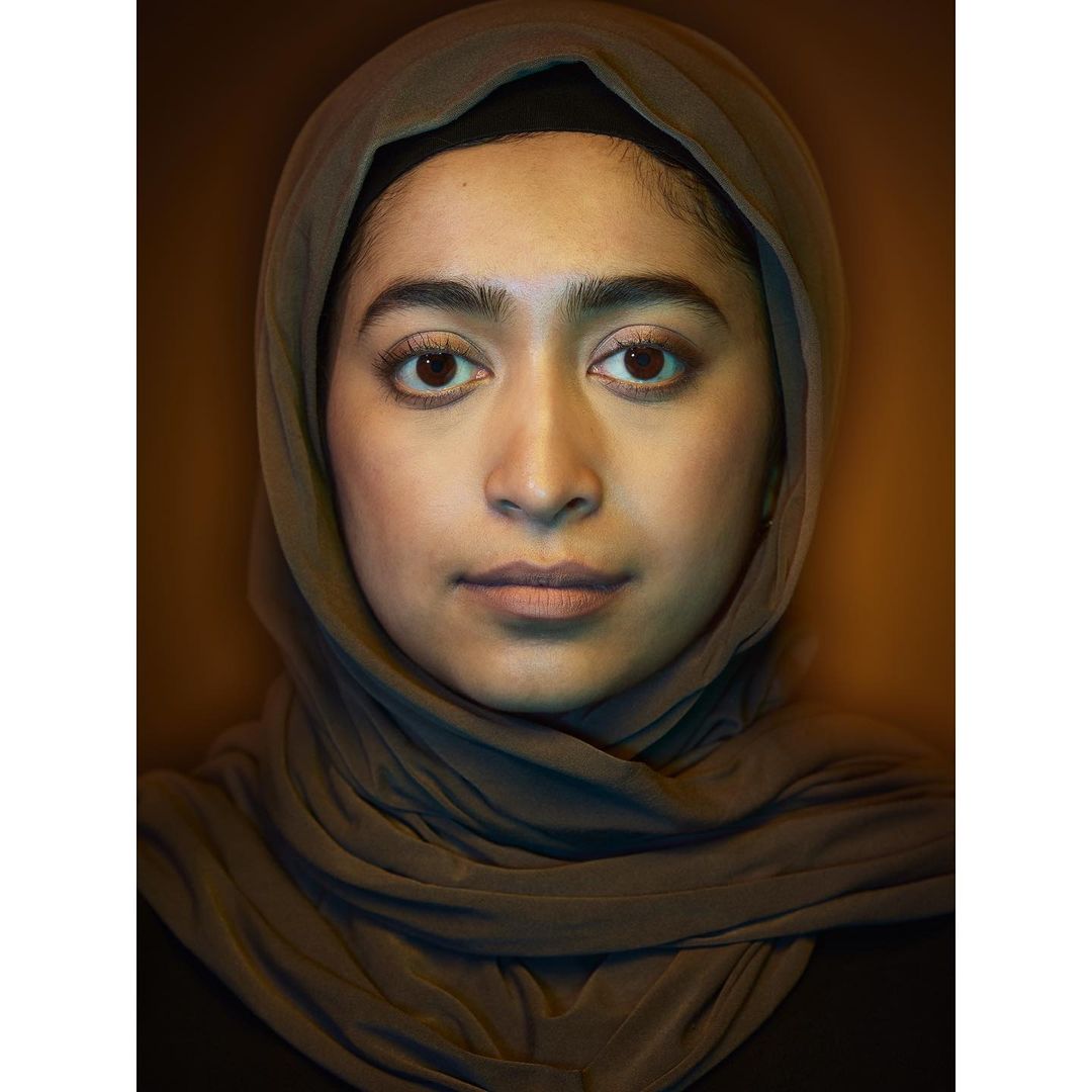 Close-up portrait of a young woman with fair skin and brown eyes, wearing a gray hijab, against a blurred brown background.