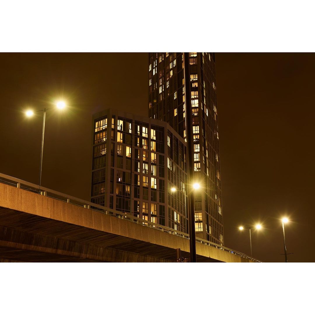 Nighttime cityscape featuring illuminated apartment buildings under a dark sky, viewed from below a bridge with street lights on.