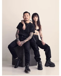 Portrait of a tattooed man and woman in black attire seated on a dark box against a light background.