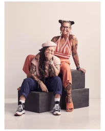 Two women with tattoos seated on dark studio blocks against a white seamless background, photographed by Dylan Collard in London.