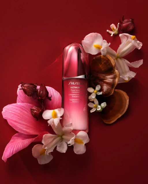 Shiseido Ultimune serum bottle surrounded by white and pink flowers and dried hibiscus on a deep red background in a beauty product shot.