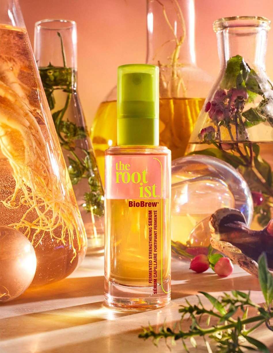 Close-up of the Rootist BioBrew serum bottle, with a green lid and a light orange liquid. Laboratory beakers in the background.