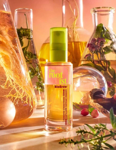 Close-up of the Rootist BioBrew serum bottle, with a green lid and a light orange liquid. Laboratory beakers in the background.