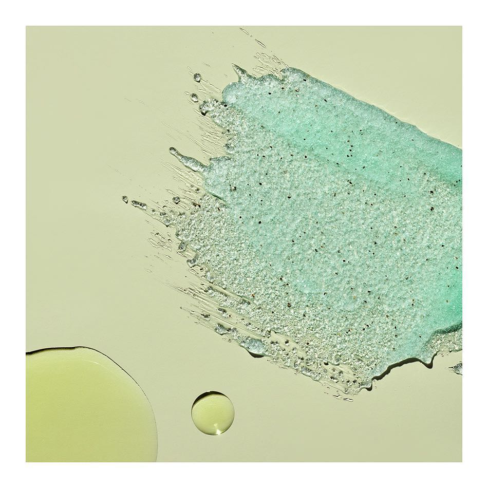 Close-up of light green cosmetic scrub and oil droplets against a beige background, suitable for beauty and skincare product promotion.