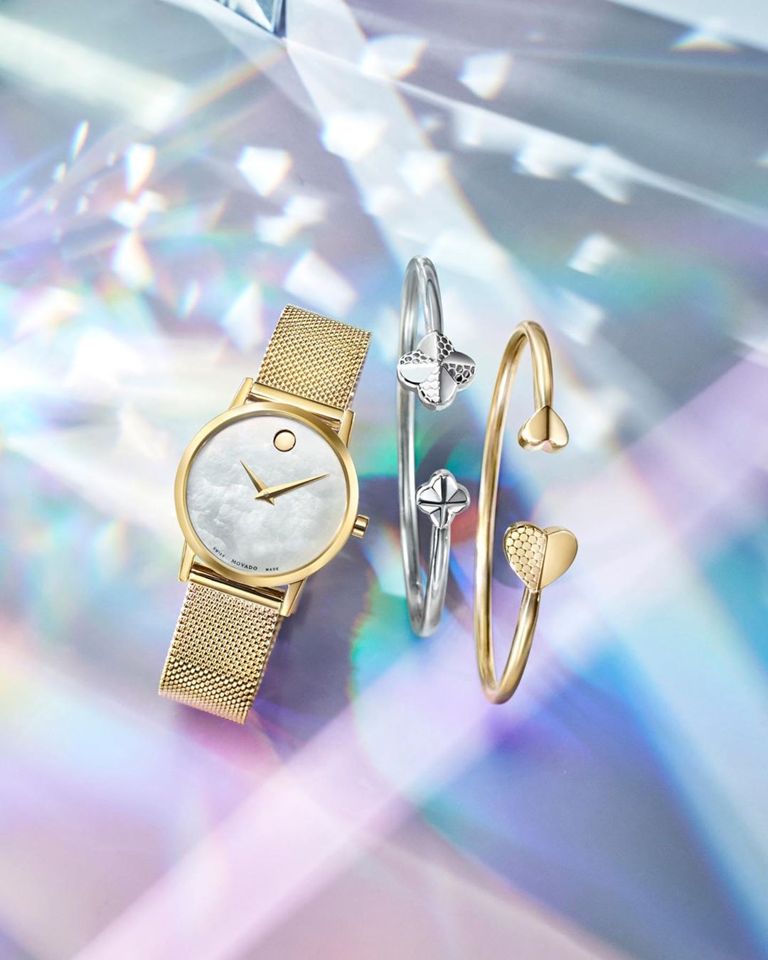 Elegant gold Movado watch with cloud-patterned face, and two gold and silver open bangle bracelets, jewelry products, on a shiny background.