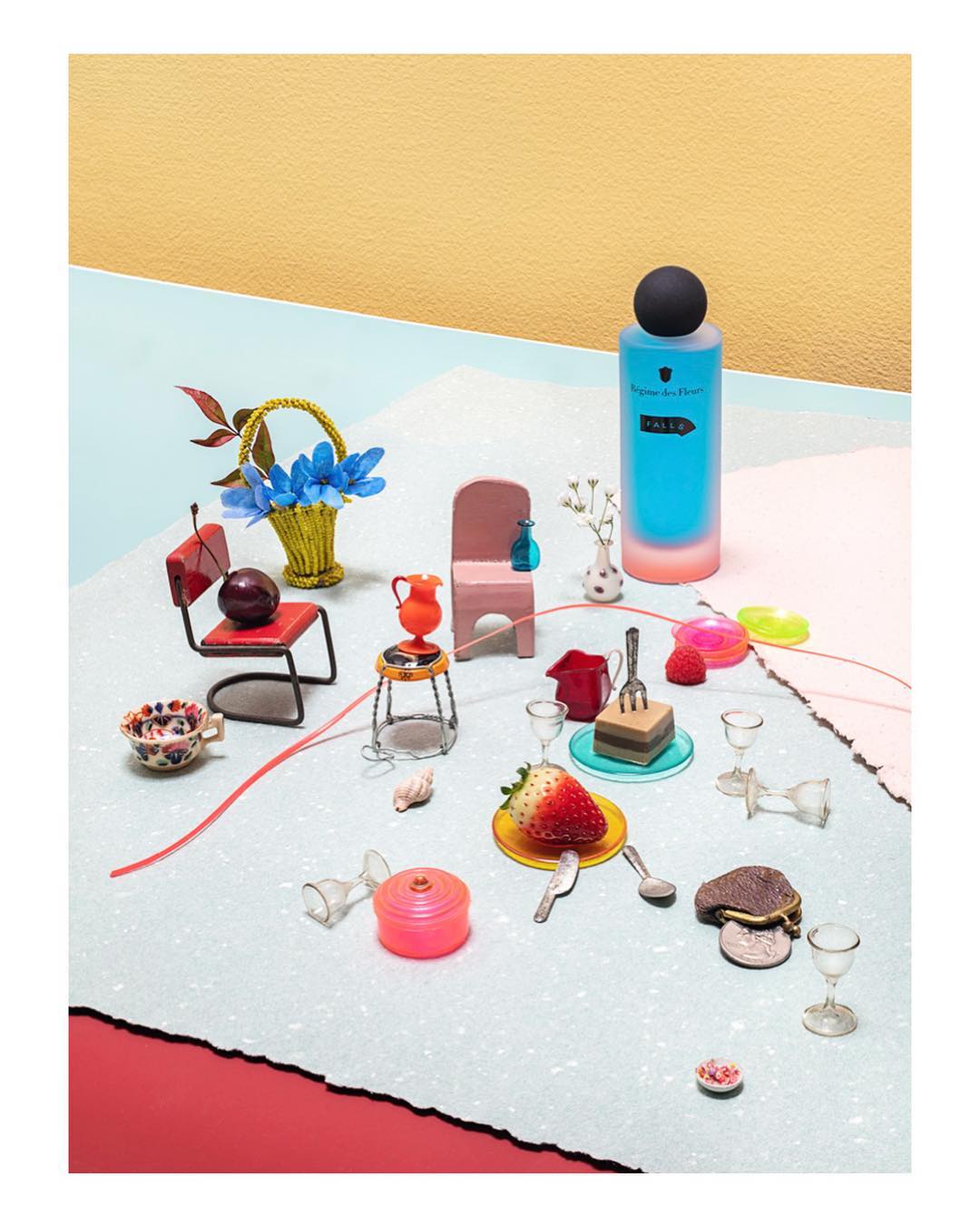 Still life composition features a perfume bottle, miniature chairs, strawberries, and various props on a light blue textured surface.