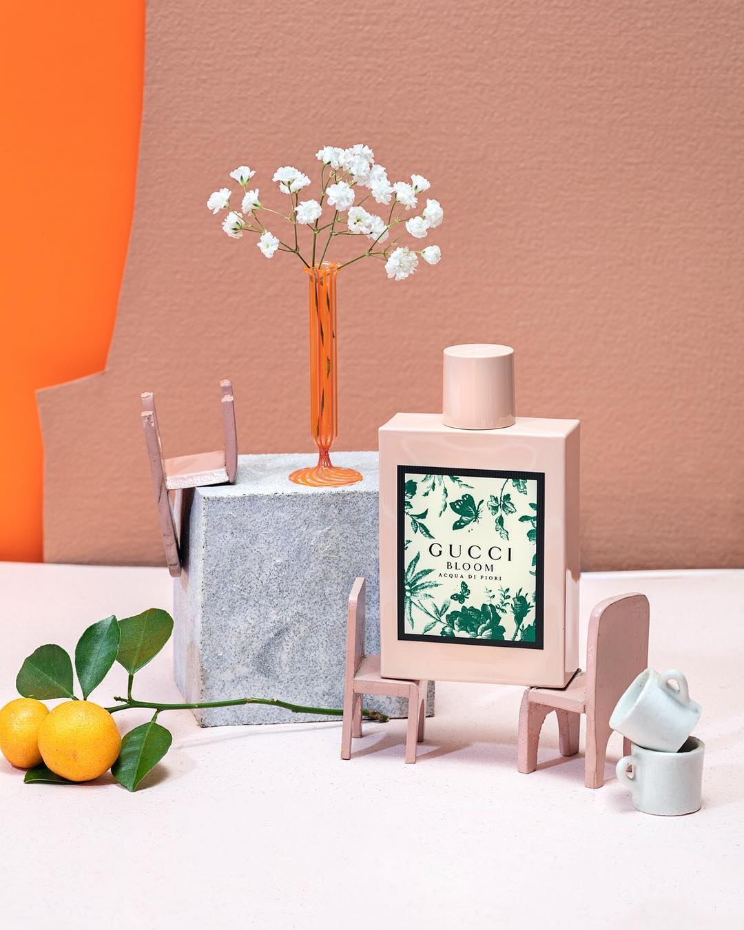 Gucci Bloom perfume bottle surrounded by miniature chairs and lemons in a creative studio product shot on a peach and orange background.