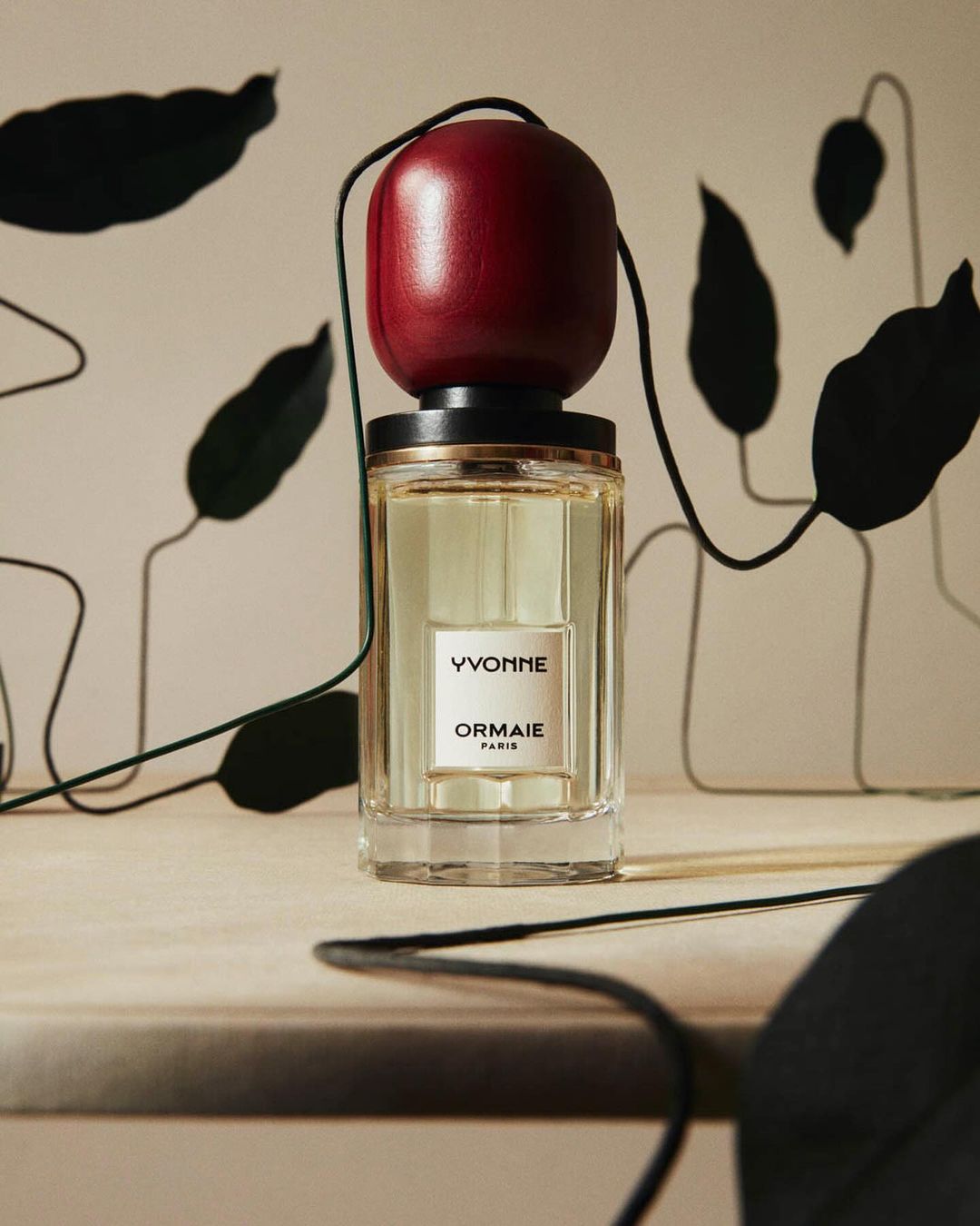 Close-up of Yvonne by Ormaie Paris perfume bottle with a red cap, set on a beige surface, against a leaf-patterned backdrop.