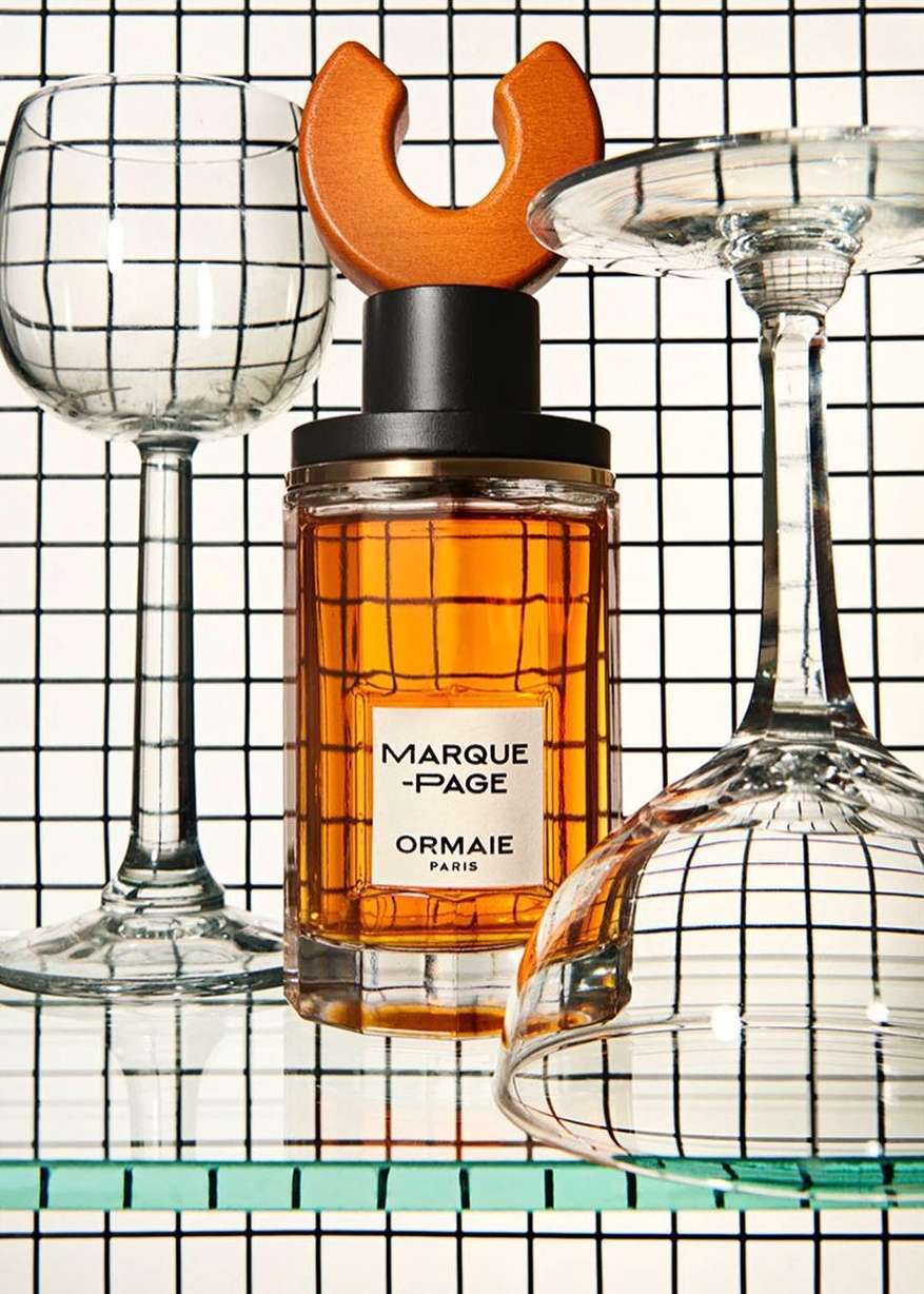 Close-up of a perfume bottle on a glass shelf with two wine glasses against a black and white checkered background, studio lighting.