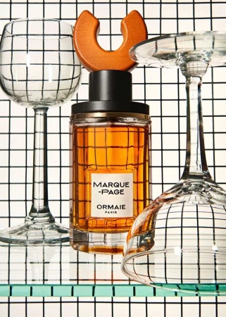 Close-up of a perfume bottle on a glass shelf with two wine glasses against a black and white checkered background, studio lighting.