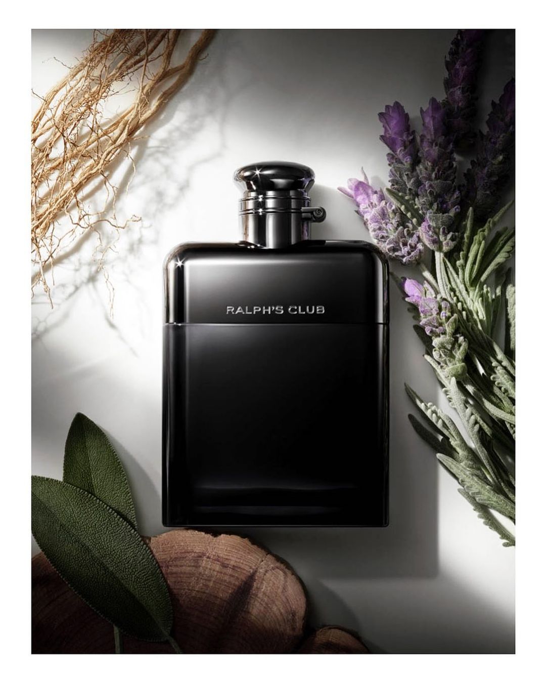 Close-up studio shot of a black Ralph's Club fragrance bottle surrounded by sage leaves, lavender, and decorative branches on white marble.