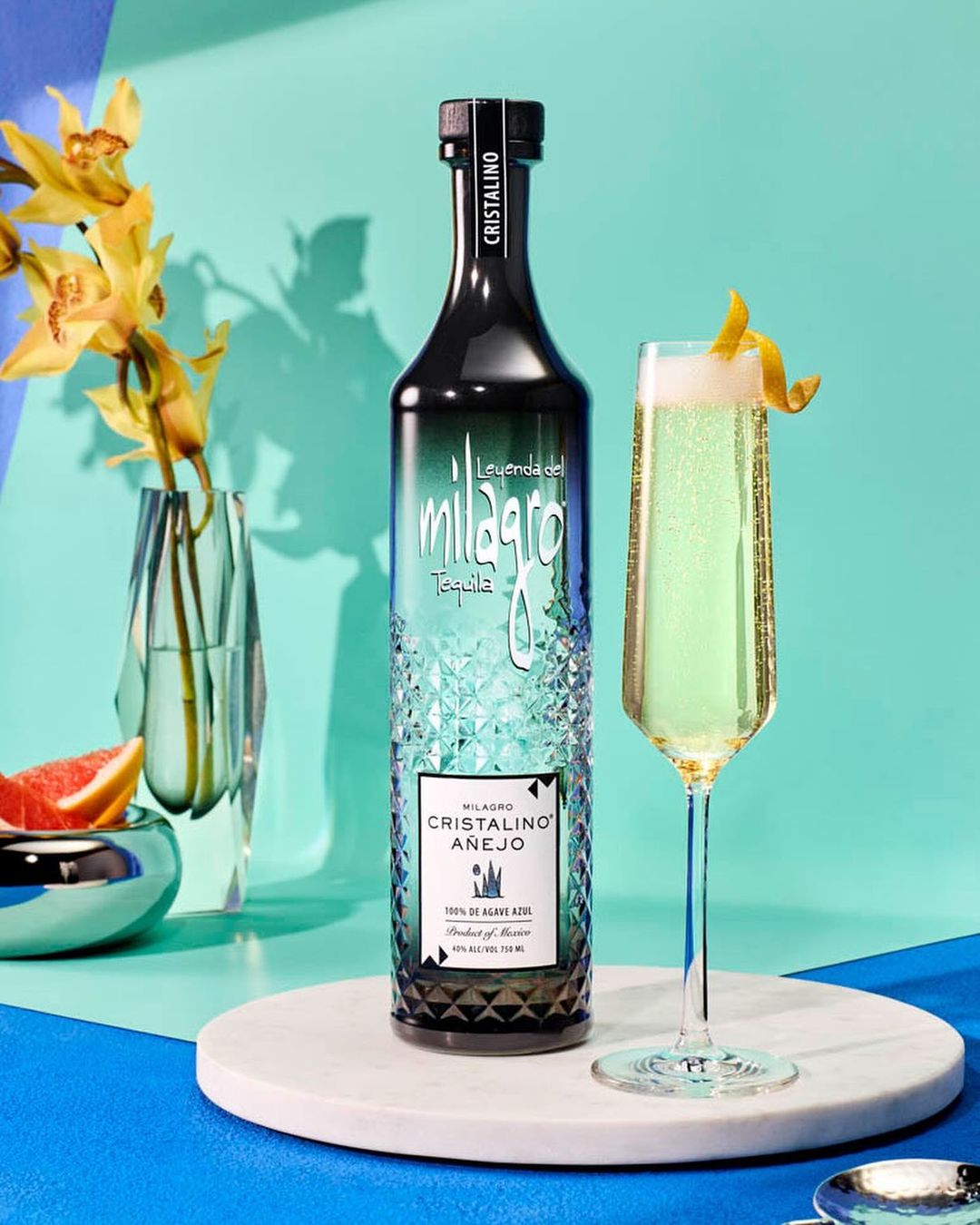 Milagro Cristalino Añejo tequila bottle next to a champagne flute filled with sparkling drink and lemon peel on a blue background.