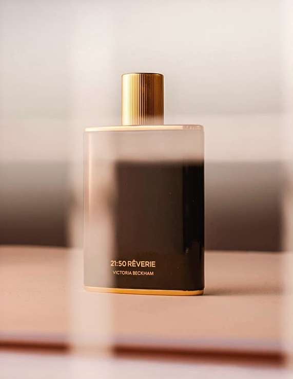 Rectangular perfume bottle with gold cap on a reflective surface, featuring '21:50 RÊVERIE VICTORIA BECKHAM' on the container.
