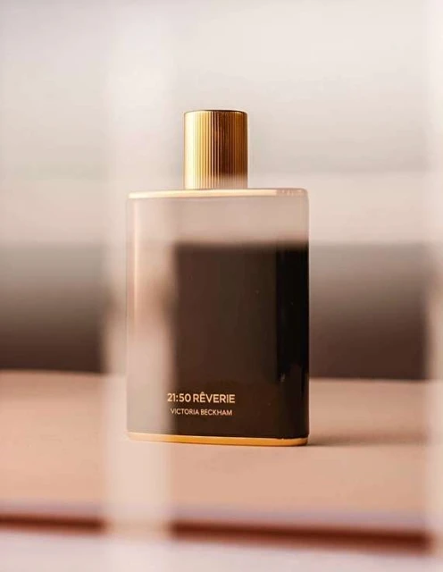 Rectangular perfume bottle with gold cap on a reflective surface, featuring '21:50 RÊVERIE VICTORIA BECKHAM' on the container.