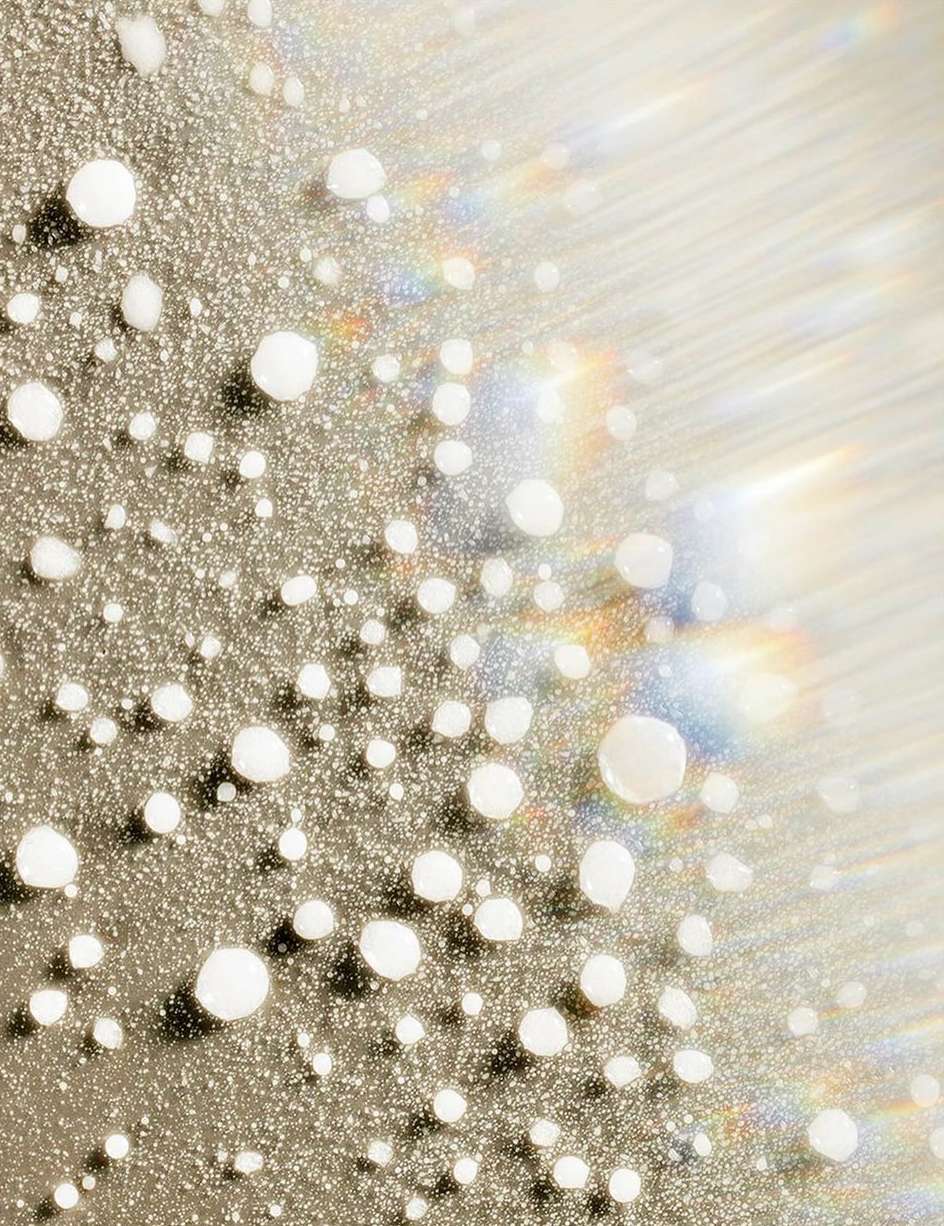 Macro shot of small white droplets on a reflective surface, creating a textured, glistening effect with soft light and rainbow flares.