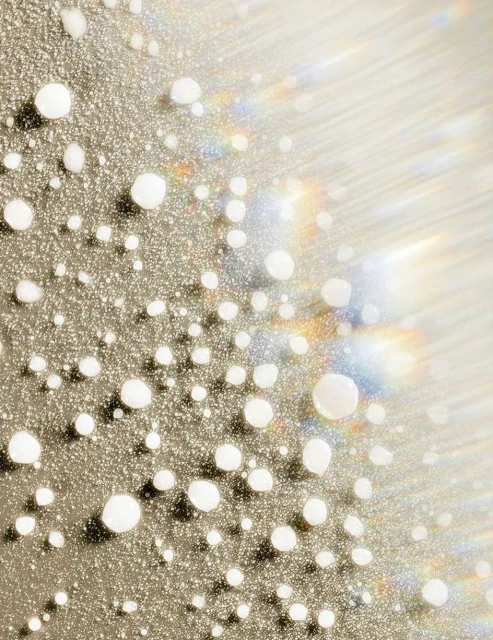 Macro shot of small white droplets on a reflective surface, creating a textured, glistening effect with soft light and rainbow flares.