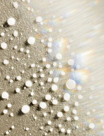 Macro shot of small white droplets on a reflective surface, creating a textured, glistening effect with soft light and rainbow flares.