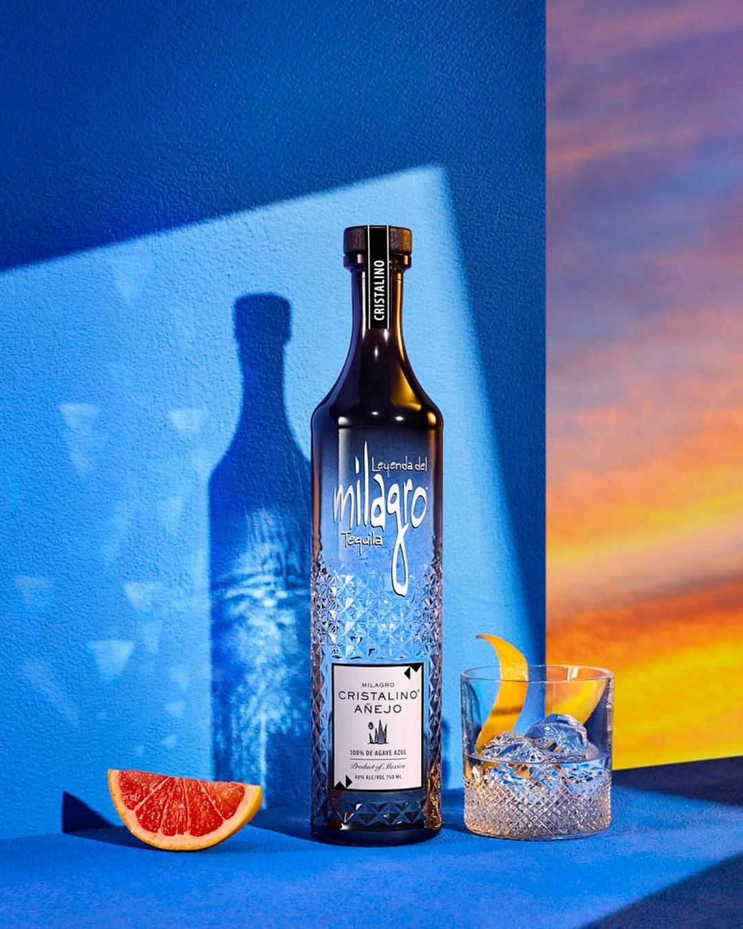 Bottle of Milagro Cristalino Añejo tequila with grapefruit slice, cocktail, blue background, studio lighting, and sunset backdrop.