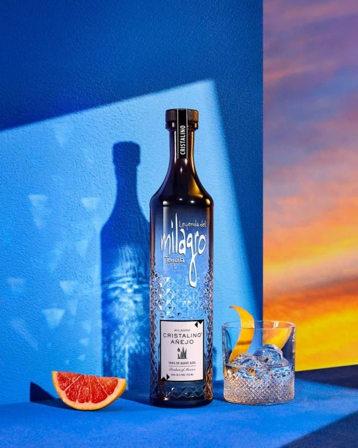 Bottle of Milagro Cristalino Añejo tequila with grapefruit slice, cocktail, blue background, studio lighting, and sunset backdrop.