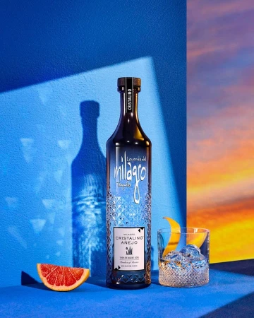 Bottle of Milagro Cristalino Añejo tequila with grapefruit slice, cocktail, blue background, studio lighting, and sunset backdrop.