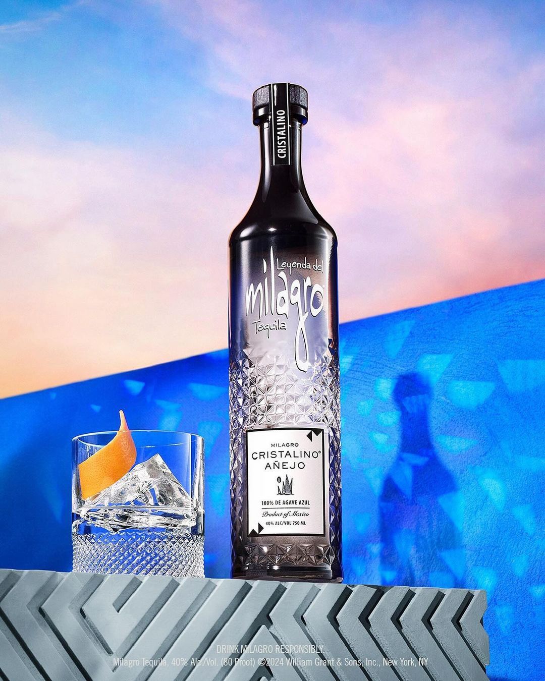 Milagro Cristalino Anejo tequila bottle, cocktail glass with ice and orange peel, against a blue and peach gradient background.