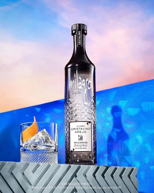 Milagro Cristalino Anejo tequila bottle, cocktail glass with ice and orange peel, against a blue and peach gradient background.