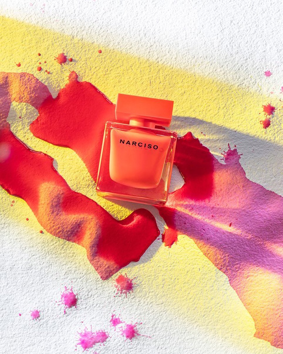 Square bottle of orange Narciso Rodriguez perfume sits on a white surface with splatters of pink and yellow liquid paint.
