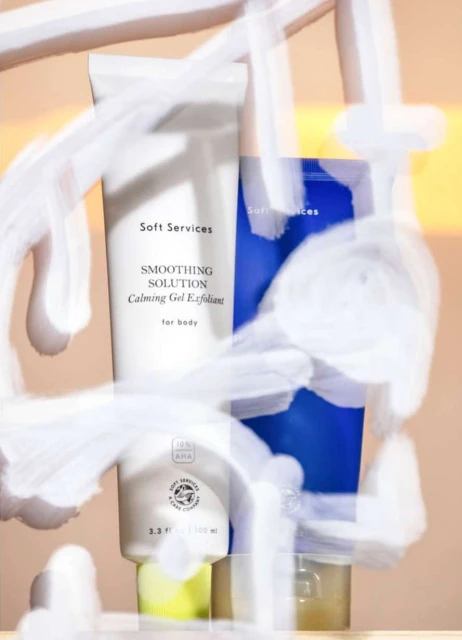 Two Soft Services skincare bottles, one white and one blue, stand side-by-side against a peach background, partially obscured by blurry white paint.
